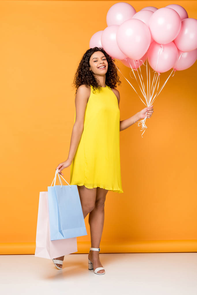 szczęśliwy African American kobieta trzyma torby na zakupy i różowe balony powietrza na pomarańczowo  - Zdjęcie, obraz