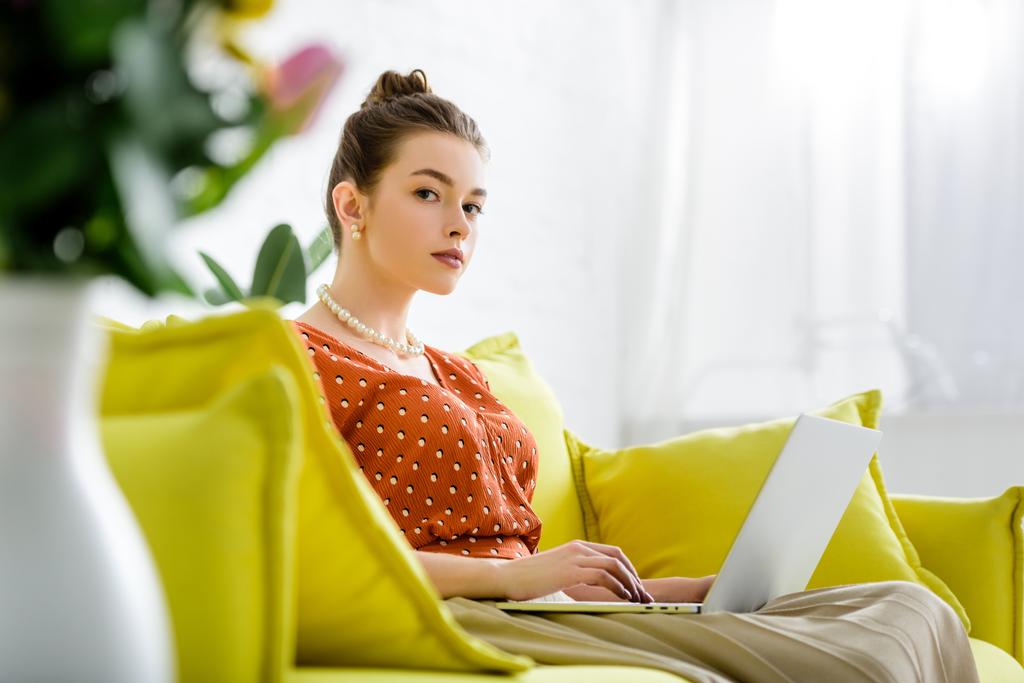 foco seletivo da mulher na moda jovem no colar de pérolas sentado no sofá amarelo e usando laptop
 - Foto, Imagem