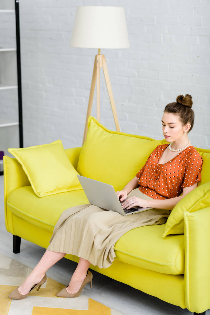 elegant young woman sitting on yellow sofa and using laptop in living room - Photo, Image