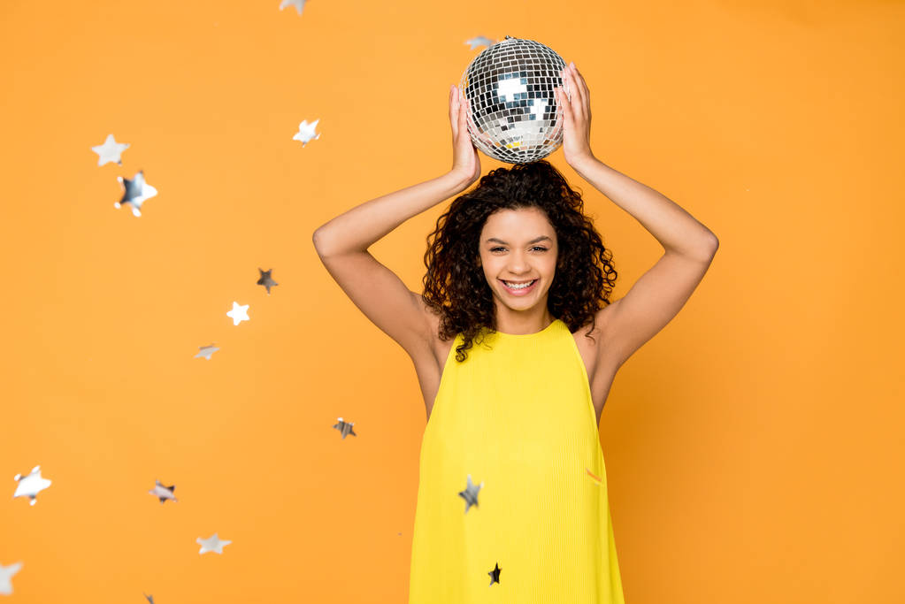 foco seletivo atraente encaracolado menina americana africana segurando bola de discoteca acima da cabeça perto de estrelas de confete brilhantes na laranja
  - Foto, Imagem