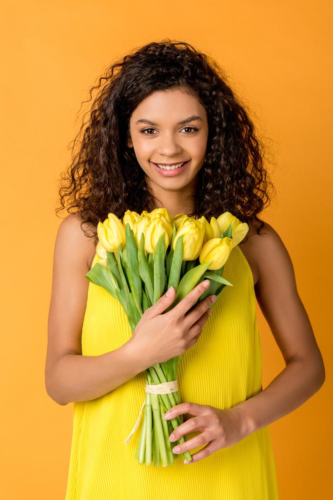 счастливая кудрявая африканская американка держит желтые тюльпаны изолированные на оранжевый
 - Фото, изображение