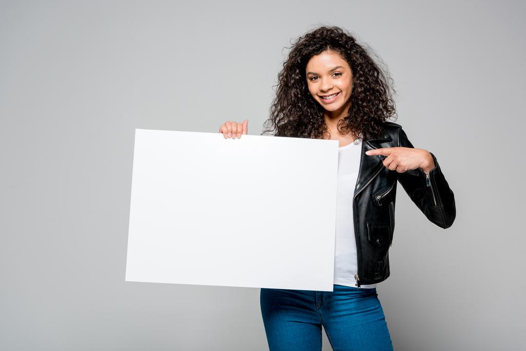 счастливая африканская американская молодая женщина, указывающая пальцем на пустой плакат, стоящая изолированная на сером
 - Фото, изображение