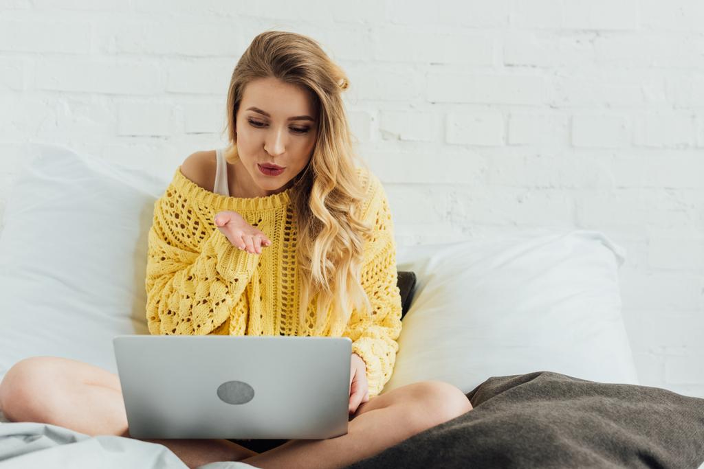 hermosa chica sentada en la cama, utilizando el ordenador portátil y soplando aire beso mientras que tiene videollamada en casa
 - Foto, Imagen