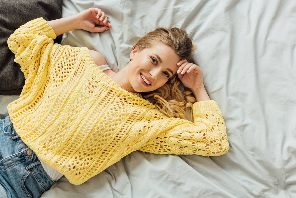 vista superior de la hermosa mujer joven en suéter de punto acostado en la cama y mirando a la cámara
 - Foto, Imagen