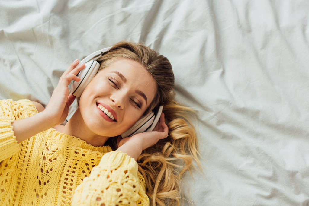 Κορυφαία θέα της πανέμορφης χαμογελαστή νεαρή γυναίκα στα ακουστικά που βρίσκονται στο κρεβάτι και ακούγοντας μουσική με χώρο αντιγραφής - Φωτογραφία, εικόνα