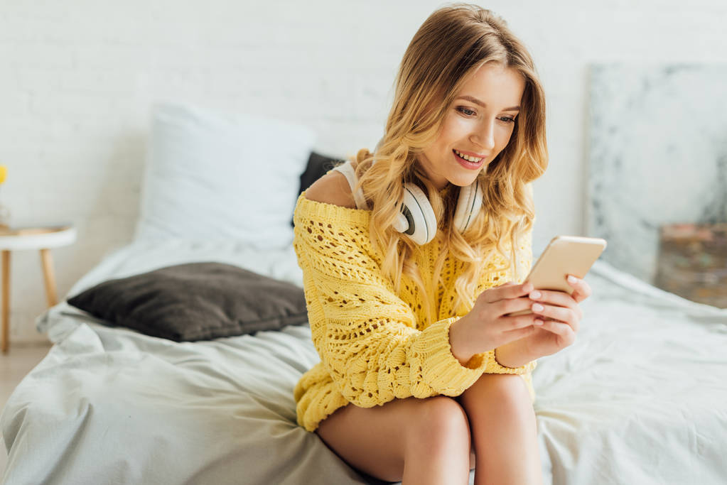 красивая девушка с наушниками, сидящая на кровати и пользующаяся смартфоном дома
 - Фото, изображение
