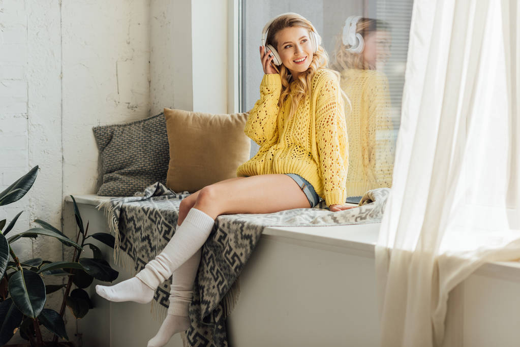 красивая улыбающаяся молодая женщина в наушниках сидит на подоконнике и слушает музыку дома
 - Фото, изображение