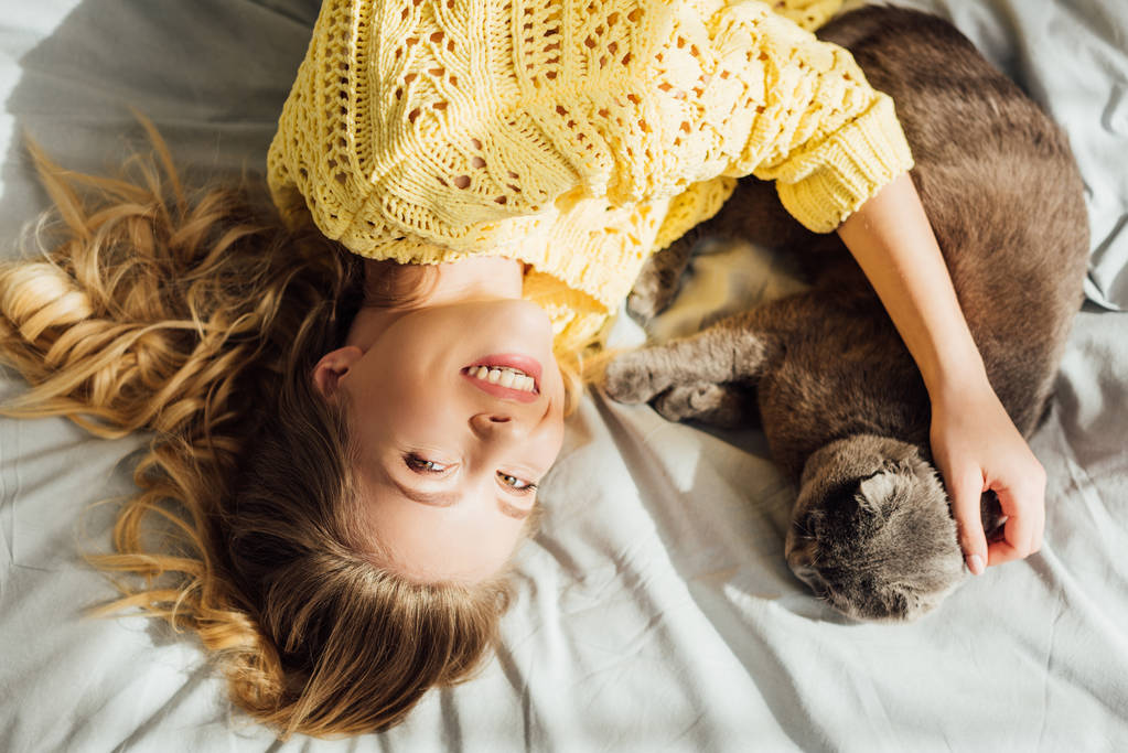 Widok z góry piękne uśmiechnięta dziewczyna głaskanie szkocki krotnie kot leżąc w łóżku w domu - Zdjęcie, obraz