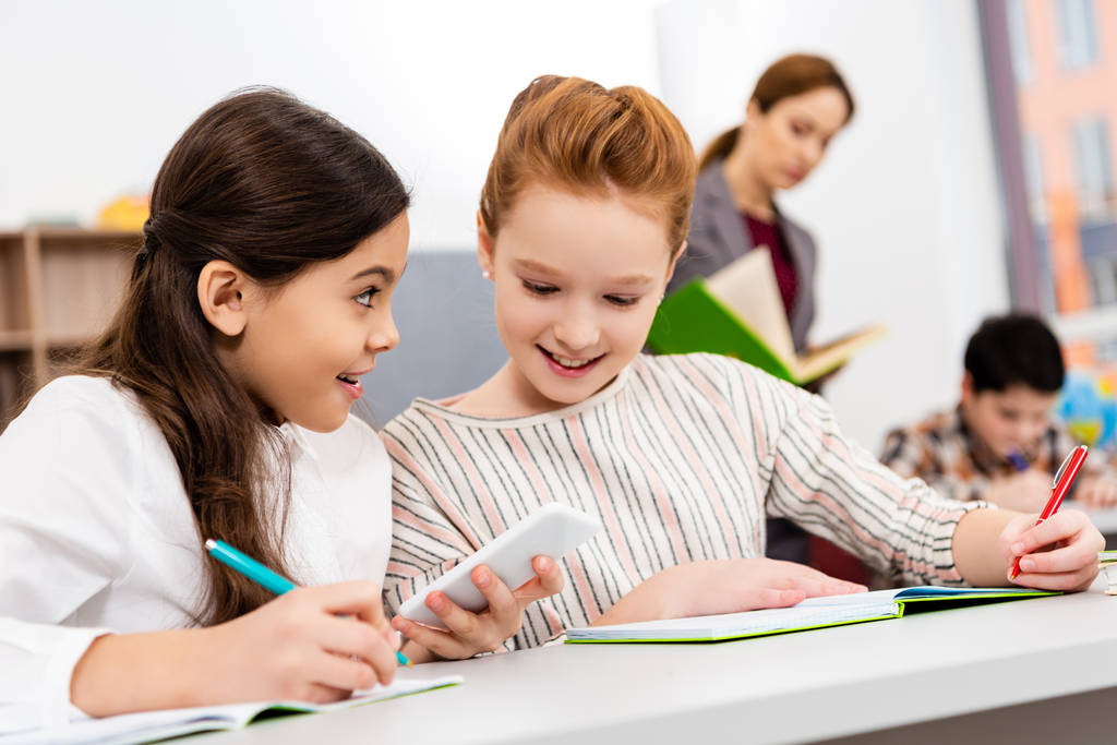 Écolières souriantes assis au bureau et utilisant un smartphone pendant les cours en classe
 - Photo, image