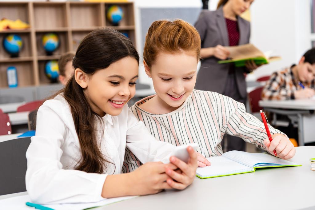 Hymyilevät koulutytöt istuvat pöydän ääressä ja käyttävät älypuhelinta oppitunnin aikana luokkahuoneessa
 - Valokuva, kuva