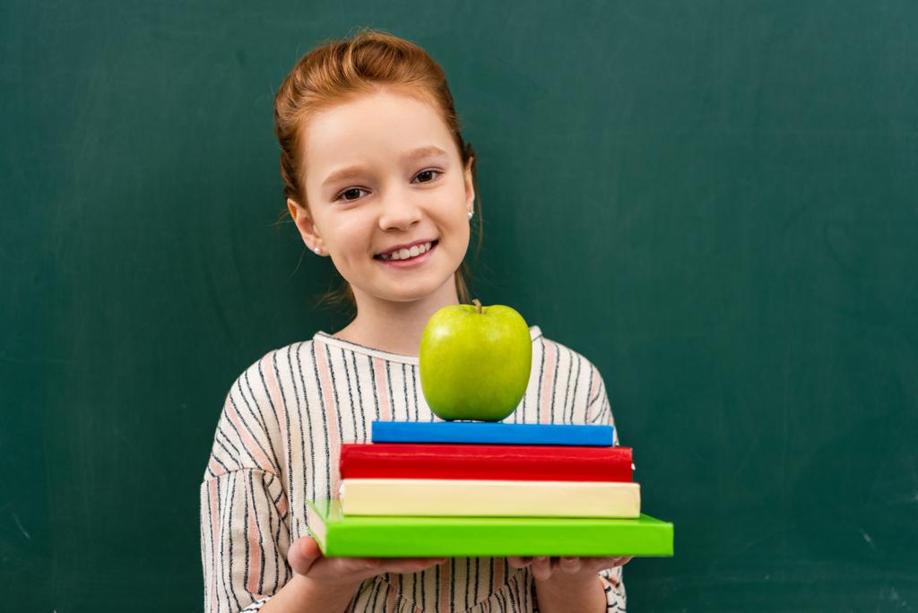 Веселая рыжая толстушка держит книги и зеленое яблоко перед школьной доской в классе
 - Фото, изображение