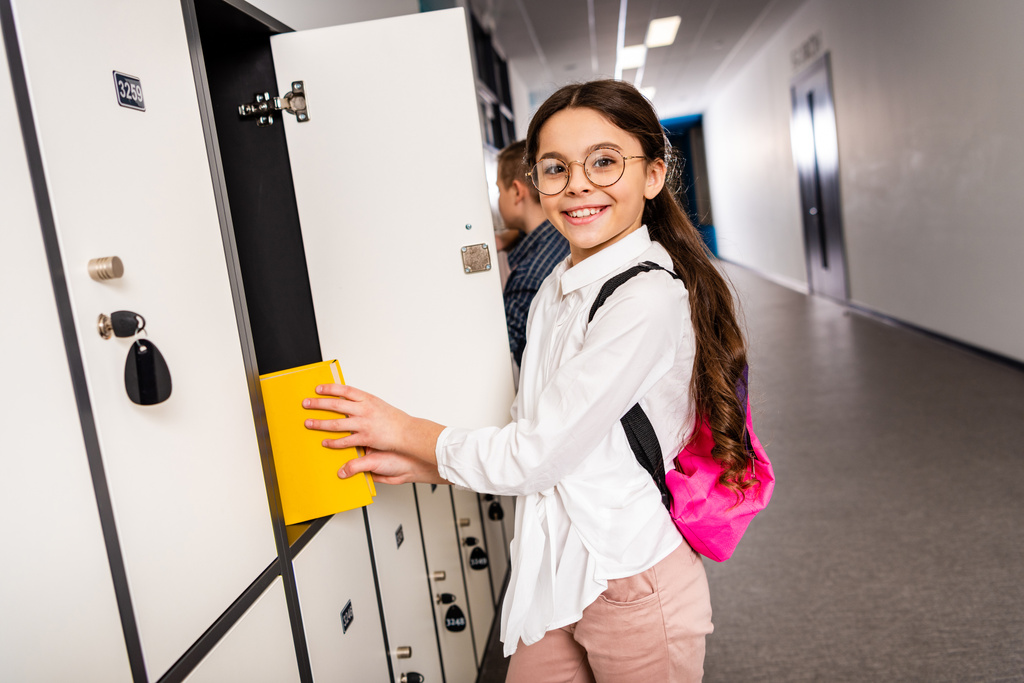 Blije school meisje in glazen putting boek in Locker tijdens de rem op school - Foto, afbeelding