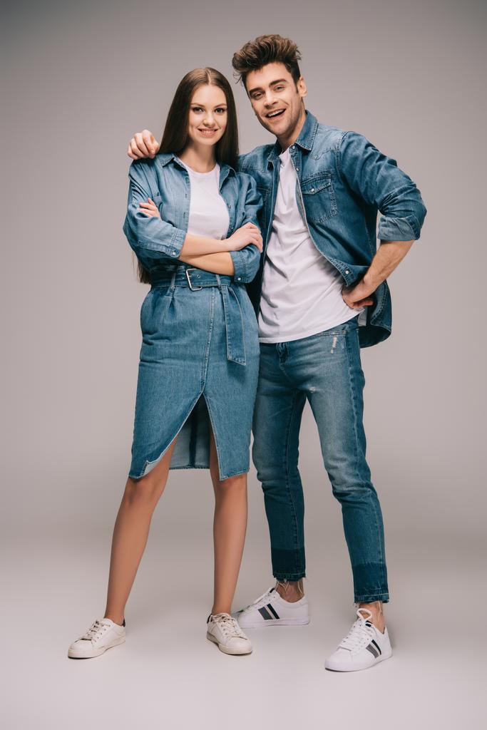 attractive girlfriend in denim dress with crossed arms and smiling boyfriend in jeans and shirt hugging and looking at camera  - Photo, Image