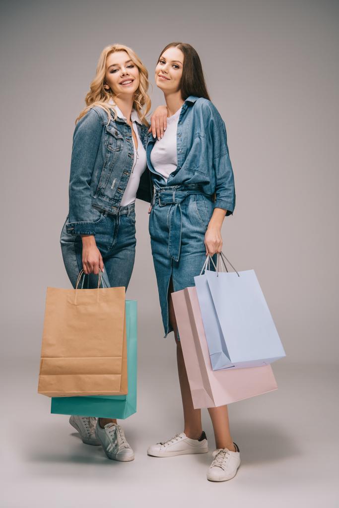 όμορφη ξανθιά και μελαχρινή γυναικών σε τζιν ρούχα, κρατώντας τσάντες για ψώνια και να βλέπουν τα φωτογραφικών μηχανών  - Φωτογραφία, εικόνα