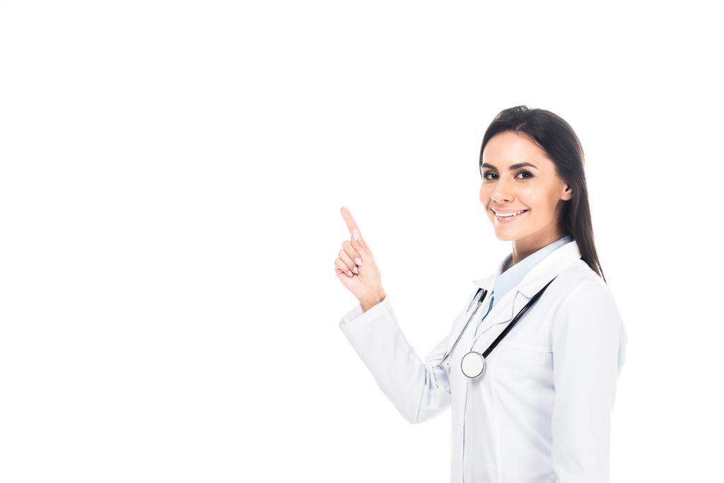 Lächelnder Arzt im weißen Mantel mit Stethoskop, das mit dem Finger auf Weiß zeigt - Foto, Bild