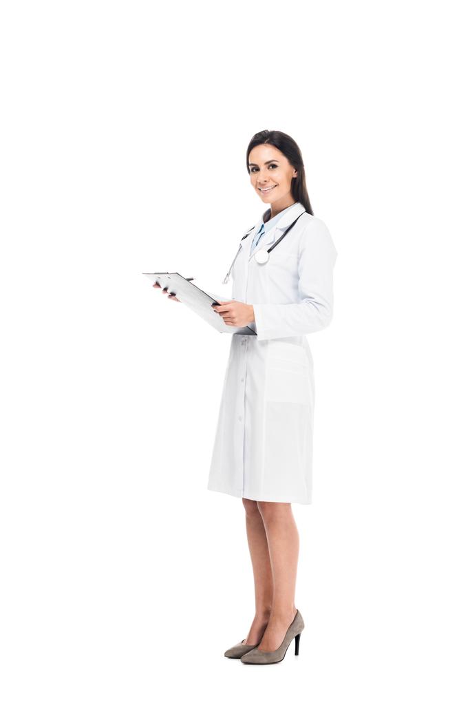 Visão de comprimento total do médico sorridente no casaco branco segurando prancheta isolada no branco
 - Foto, Imagem