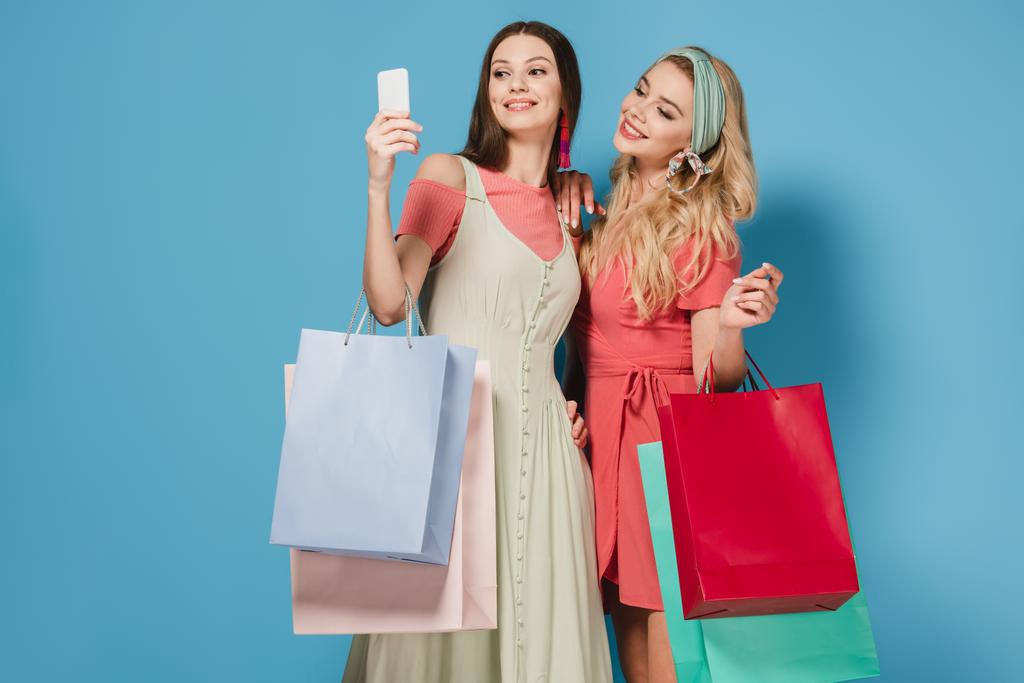 smiling brunette and blonde women in dresses holding shopping bags and taking selfie  - Photo, Image