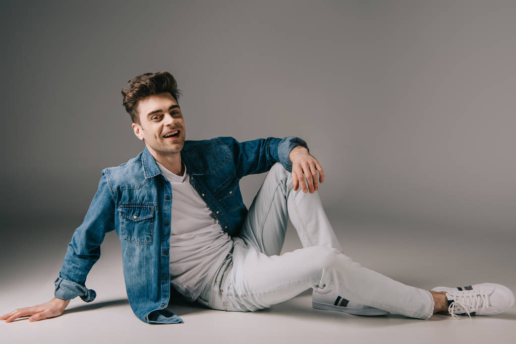smiling handsome man in denim shirt and jeans sitting on floor and looking at camera  - Photo, Image