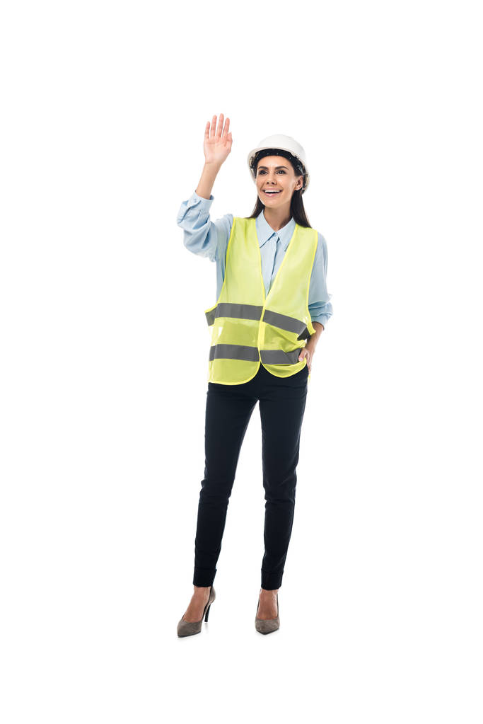 Volledige lengte weergave van lachende ingenieur in veiligheid vest zwaaien hand geïsoleerd op wit - Foto, afbeelding