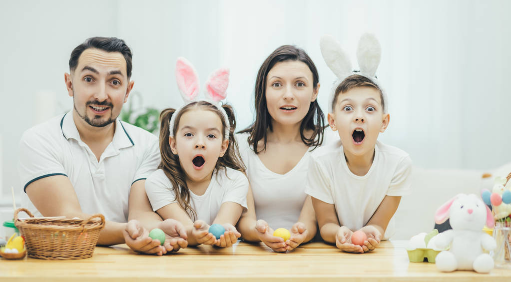 Familia inspirada en camisetas blancas sentadas juntas en la mesa, sosteniendo huevos de Pascua de colores en sus manos, mirando con entusiasmo a la cámara con expresiones de cara sorprendida. Sus bocas están abiertas
. - Foto, Imagen