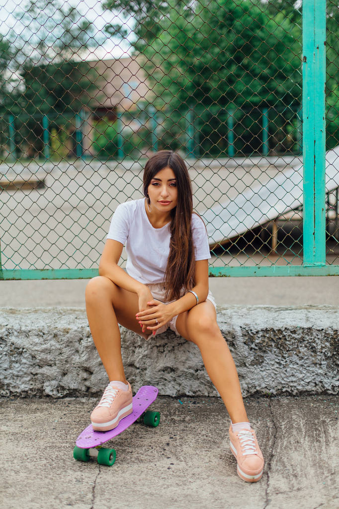 Sommer-Lifestyle-Bild des trendigen hübschen jungen Mädchens, das neben dem Skateboard-Wagen mit seinem Plastik-Skateboard sitzt. - Foto, Bild