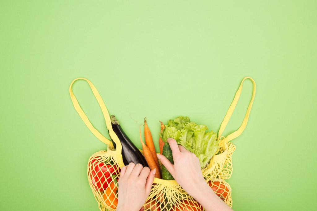 partial view of woman taking fresh cucumber out of yellow string bag with whole vegetables on light green surface - Photo, Image
