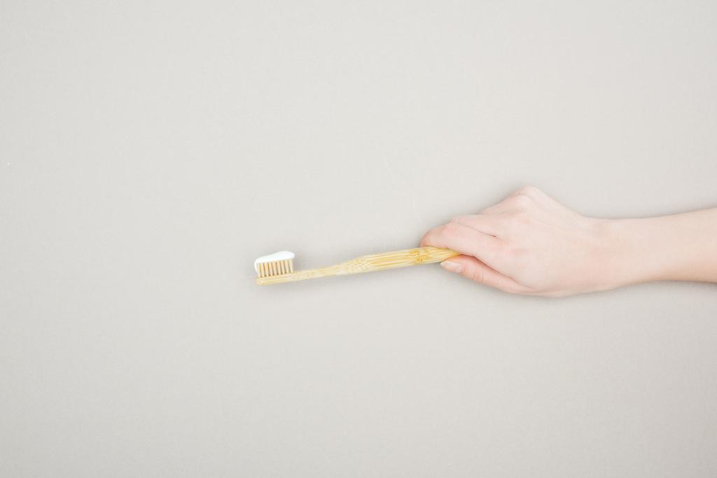 oříznutý pohled ženy držící bambusový zubní kartáček s zubní pastí na šedém pozadí - Fotografie, Obrázek