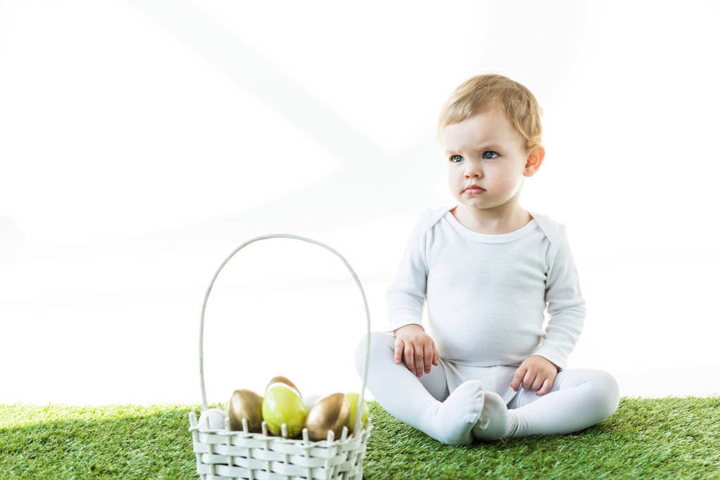 niedliches Baby sitzt auf grünem Gras in der Nähe von Strohkorb mit bunten östlichen Eiern isoliert auf weiß - Foto, Bild