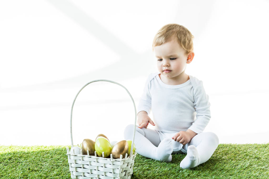 niedliches Kind sitzt in der Nähe von Strohkorb mit bunten Ostereiern isoliert auf weiß - Foto, Bild