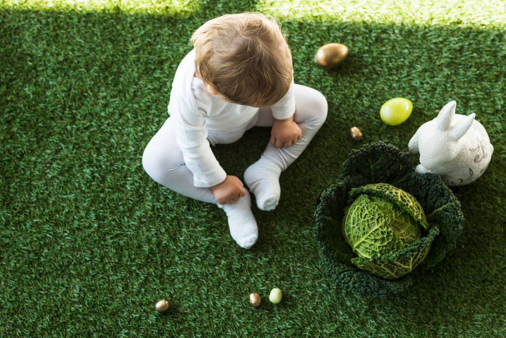 εναέρια θέα του χαριτωμένο παιδί κάθεται σε πράσινο γρασίδι κοντά Πασχαλινά αυγά, διακοσμητικό κουνέλι και λάχανο Savoy - Φωτογραφία, εικόνα