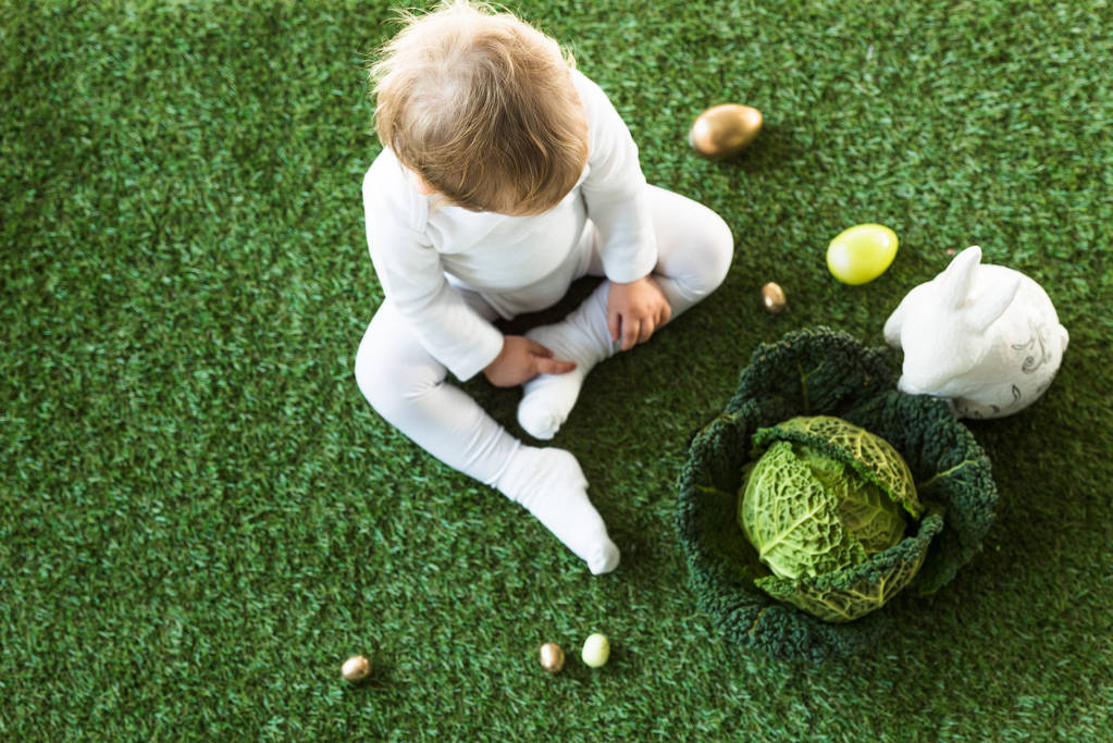 top view of blonde baby sitting on green grass near Easter eggs, decorative rabbit and savoy cabbage - Photo, Image