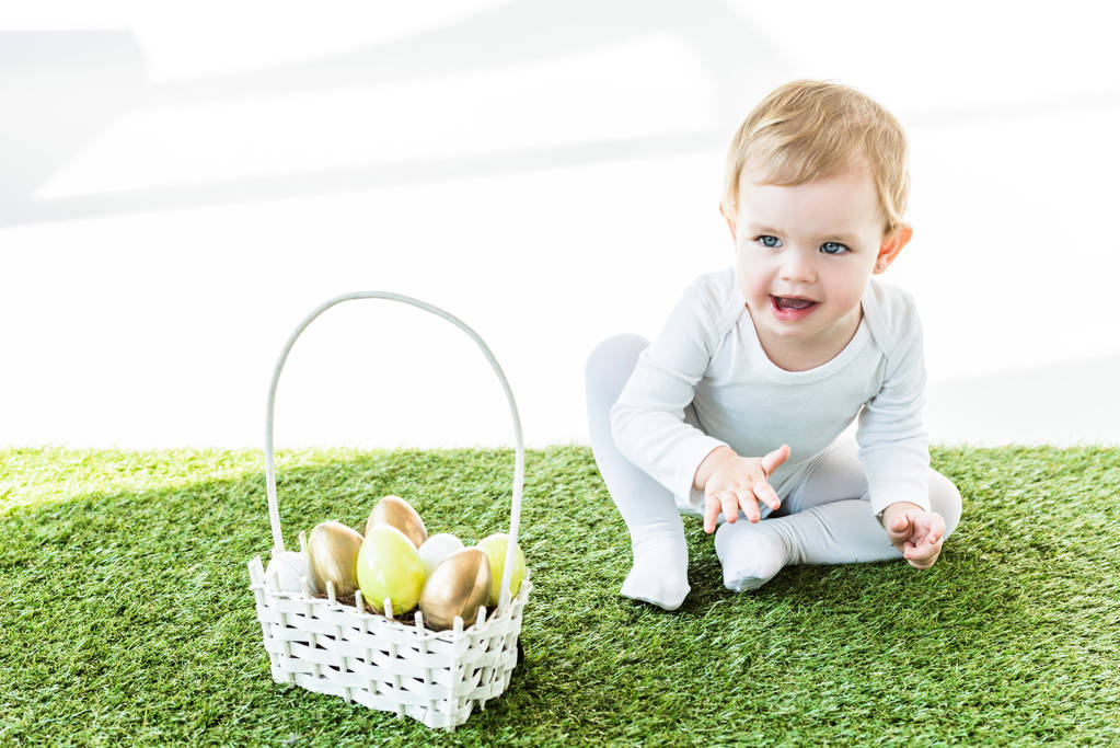 glückliches Kind sitzt neben Strohkorb mit bunten Ostereiern isoliert auf weiß - Foto, Bild