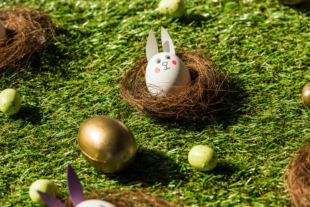 Huevos de Pascua y huevo de gallina con cara divertida de conejo y orejas de papel sobre hierba verde
   - Foto, imagen