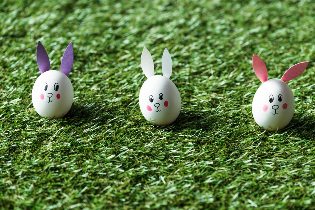 緑の草の表面に描かれたウサギの顔と紙の耳と鶏の卵の選択的焦点 - 写真・画像