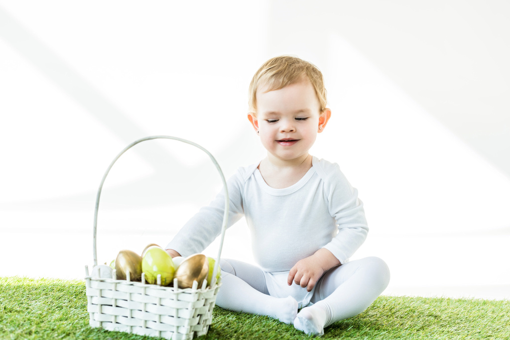 bebê sorrindo bonito sentado perto da cesta de palha com ovos de Páscoa isolados em branco
 - Foto, Imagem