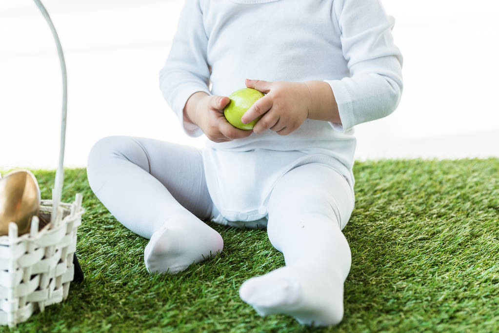 partial view of baby sitting on green grass and holding yellow chicken egg isolated on white - Photo, Image