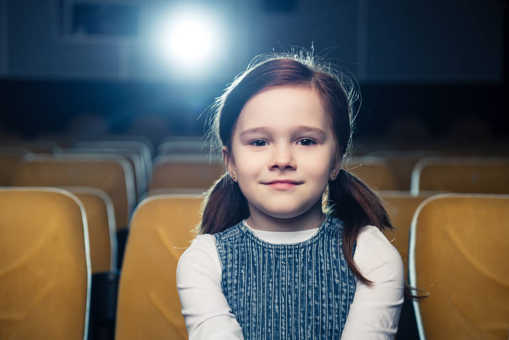 χαριτωμένο μελαχρινή παιδί κάθεται στο σινεμά και να βλέπουν φωτογραφική μηχανή  - Φωτογραφία, εικόνα