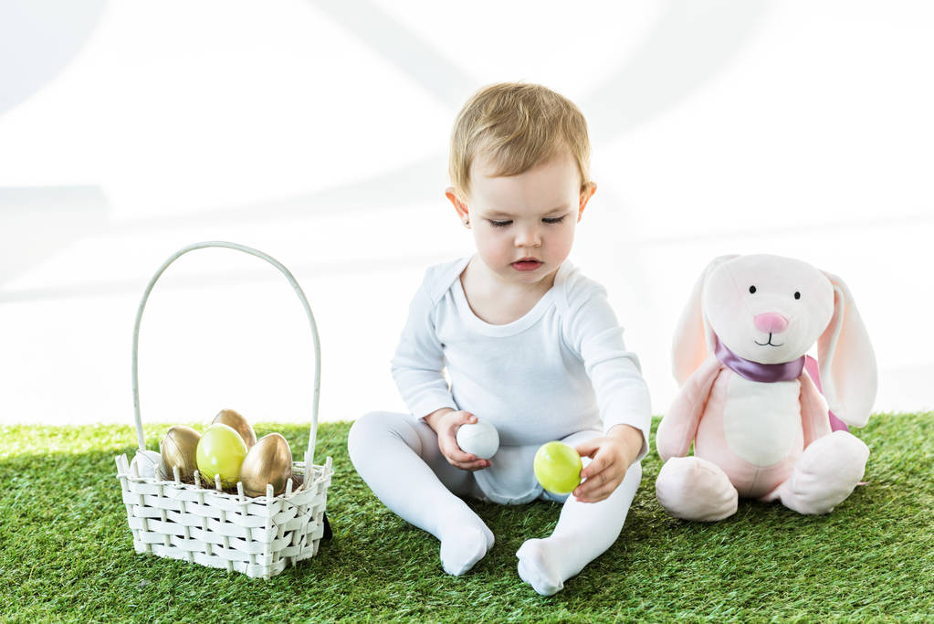 entzückendes Kind mit bunten Hühnereiern in der Nähe von Strohkorb und Spielzeugkaninchen isoliert auf weiß  - Foto, Bild