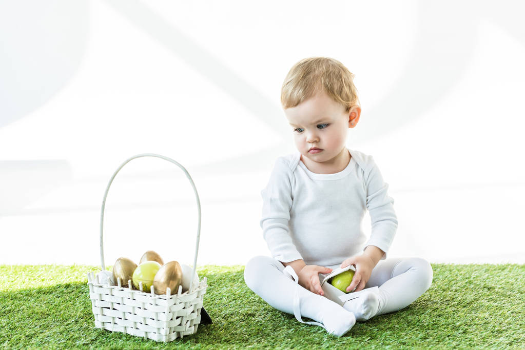 очаровательный ребенок сидит на зеленой траве рядом желтые и золотые пасхальные яйца изолированы на белом
 - Фото, изображение