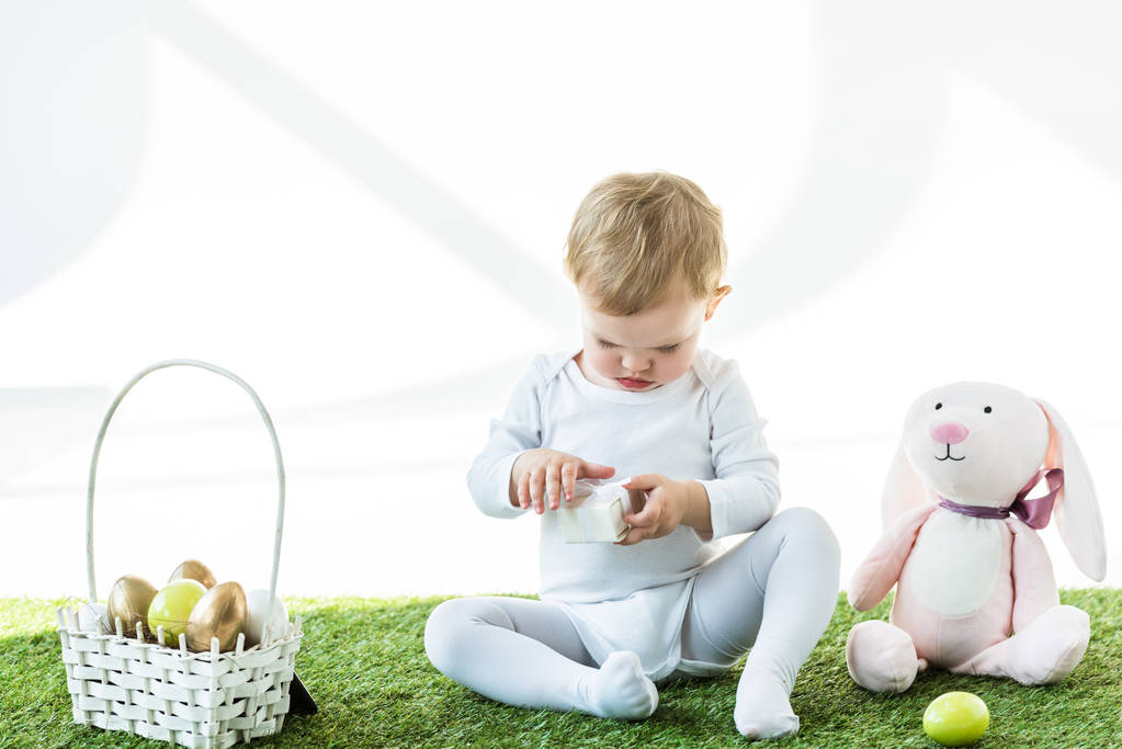 entzückendes Kind hält weiße Schachtel, während es auf grünem Gras in der Nähe von Spielzeugkaninchen und Strohkorb mit Ostereiern sitzt, isoliert auf weißem Grund - Foto, Bild
