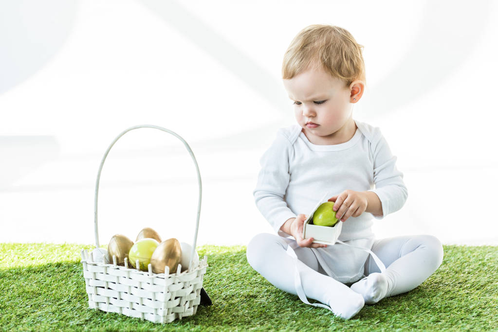 cute child holding box with yellow egg and looking at straw basket with Easter eggs isolated on white - Photo, Image
