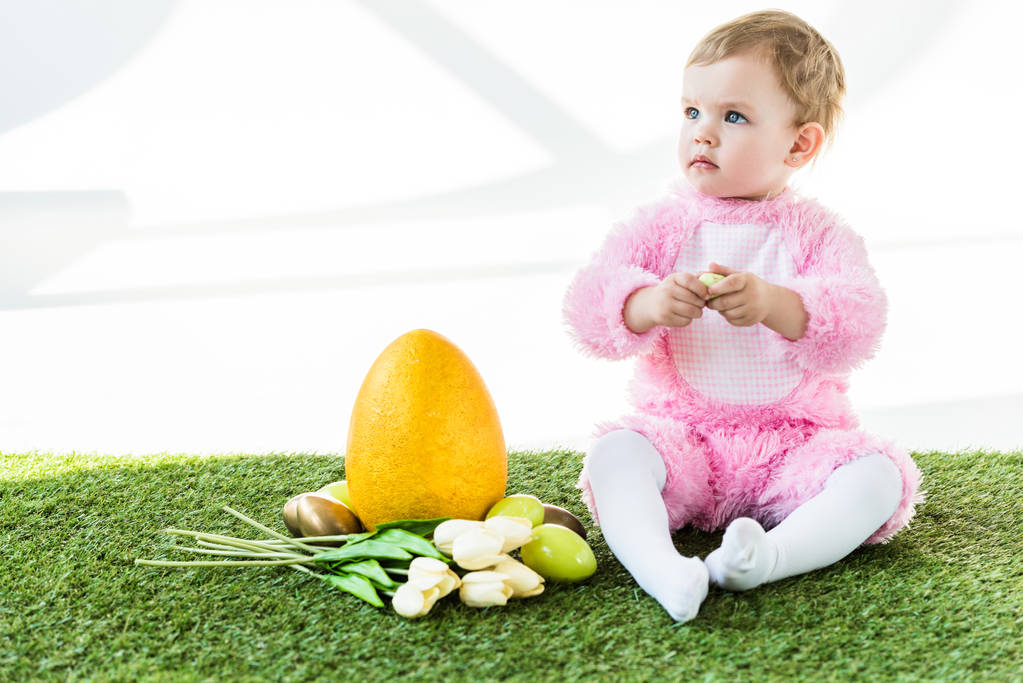 niedliches Baby im rosafarbenen flauschigen Kostüm sitzt neben gelben Straußenei, bunten Hühnereiern und Tulpen isoliert auf weiß - Foto, Bild