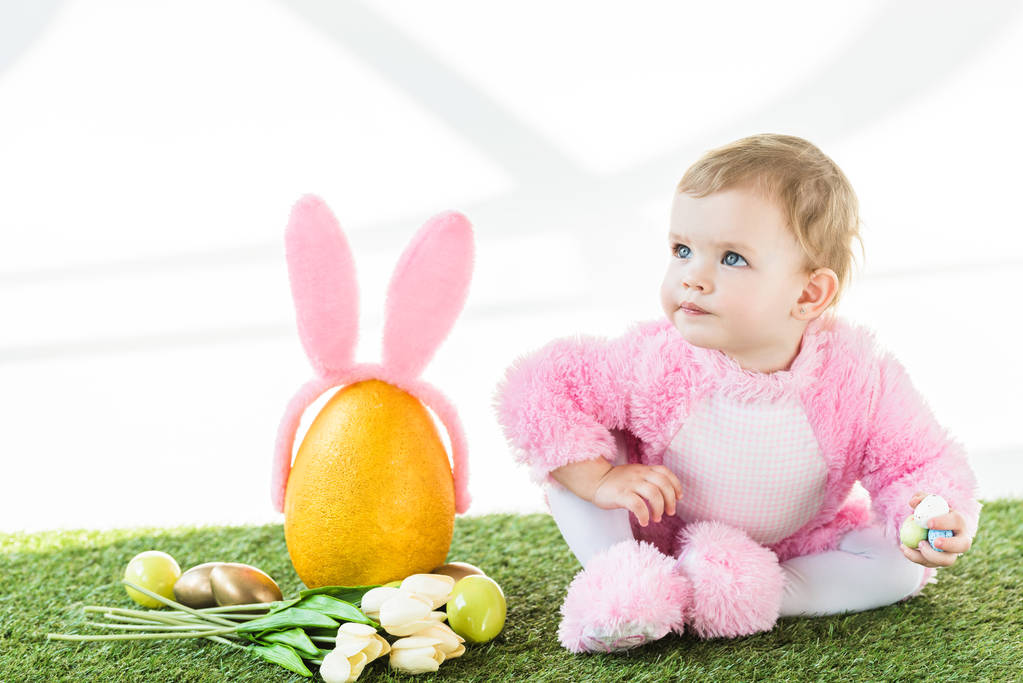 adorable niño en traje mullido rosa sentado cerca de huevo de avestruz amarillo con diadema de orejas de conejo, coloridos huevos de Pascua y tulipanes aislados en blanco
 - Foto, Imagen