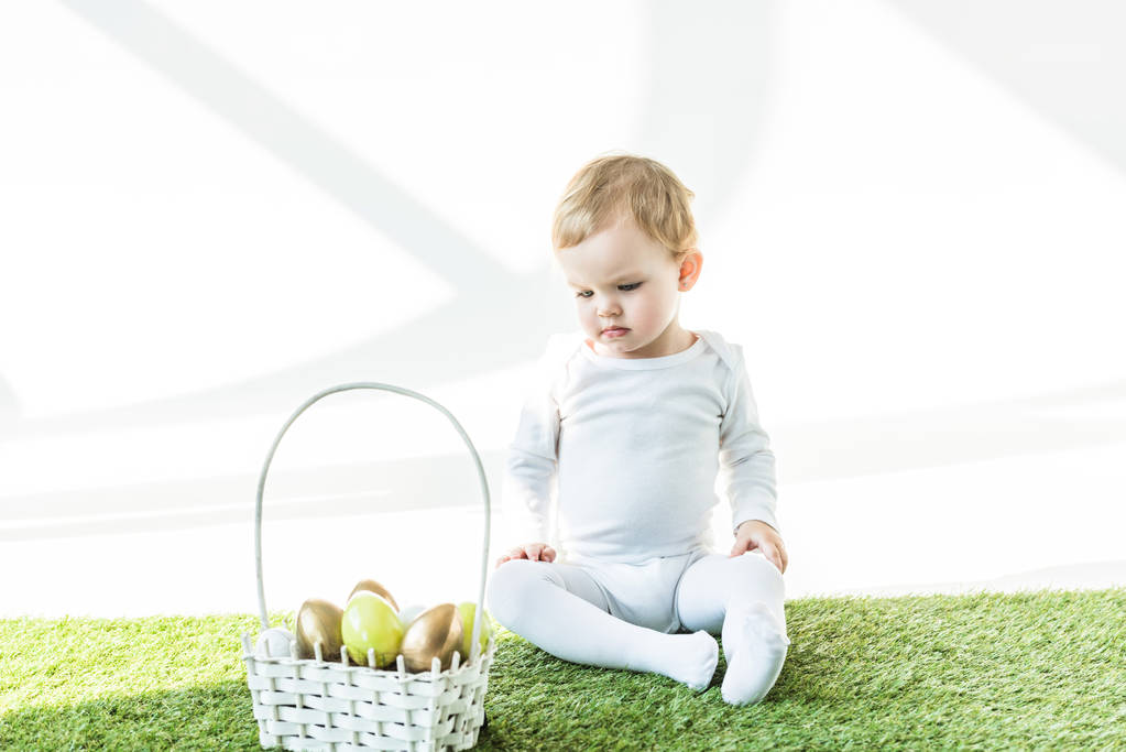 cute child sitting on green grass near straw basket with colorful Easter eggs isolated on white  - Photo, Image