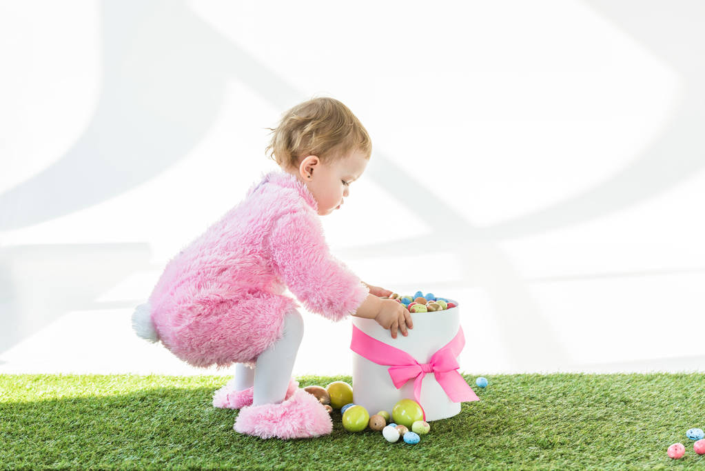 lindo niño en traje mullido rosa de pie cerca de la caja con arco rosa y coloridos huevos de Pascua aislados en blanco
 - Foto, Imagen