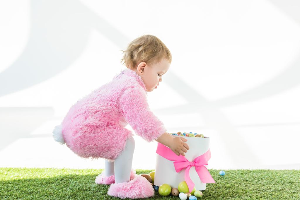 criança adorável no traje fofo rosa que está perto da caixa com arco rosa e ovos de Páscoa coloridos isolados no branco
 - Foto, Imagem