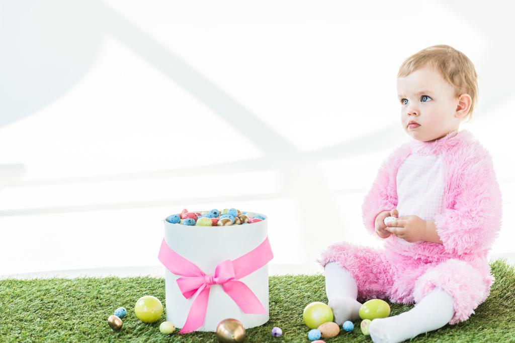 αξιολάτρευτο παιδί σε ροζ χνουδωτή φορεσιά καθισμένος κοντά στο κουτί με ροζ τόξο και πολύχρωμα Πασχαλινά αυγά απομονωμένα σε λευκό - Φωτογραφία, εικόνα