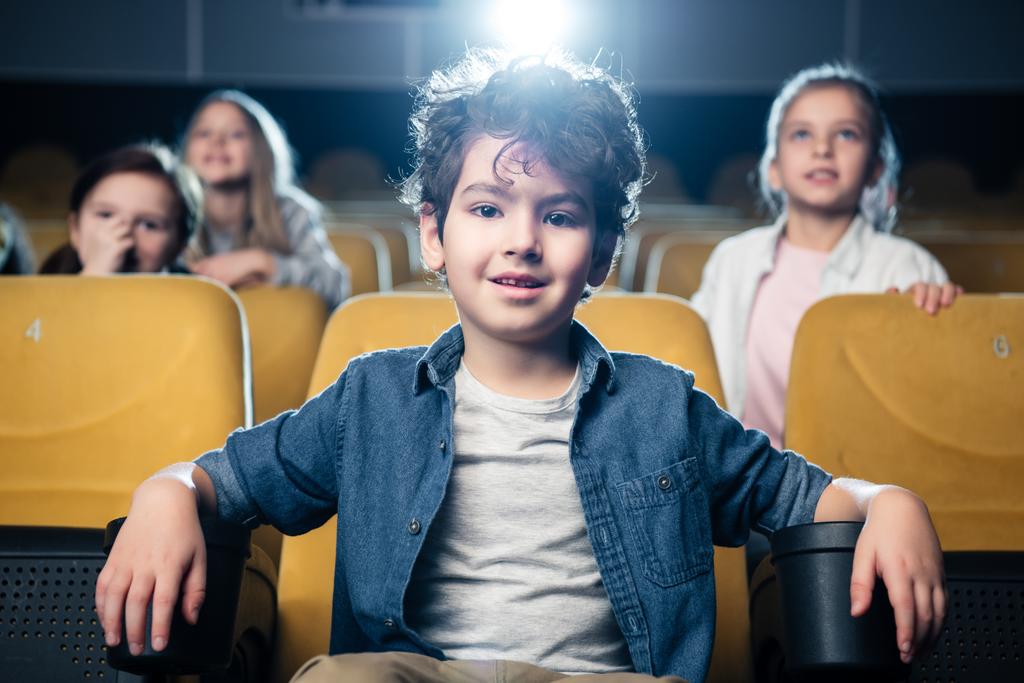 chłopiec cute mieszane rasy patrząc na kamery, siedząc w kinie wraz z przyjaciółmi - Zdjęcie, obraz
