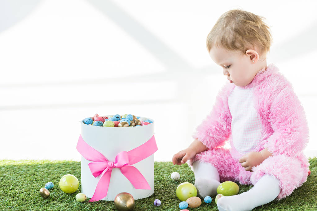 adorabile bambino in costume rosa soffice seduto su erba verde vicino alla scatola con uova di Pasqua colorate isolate su bianco
 - Foto, immagini
