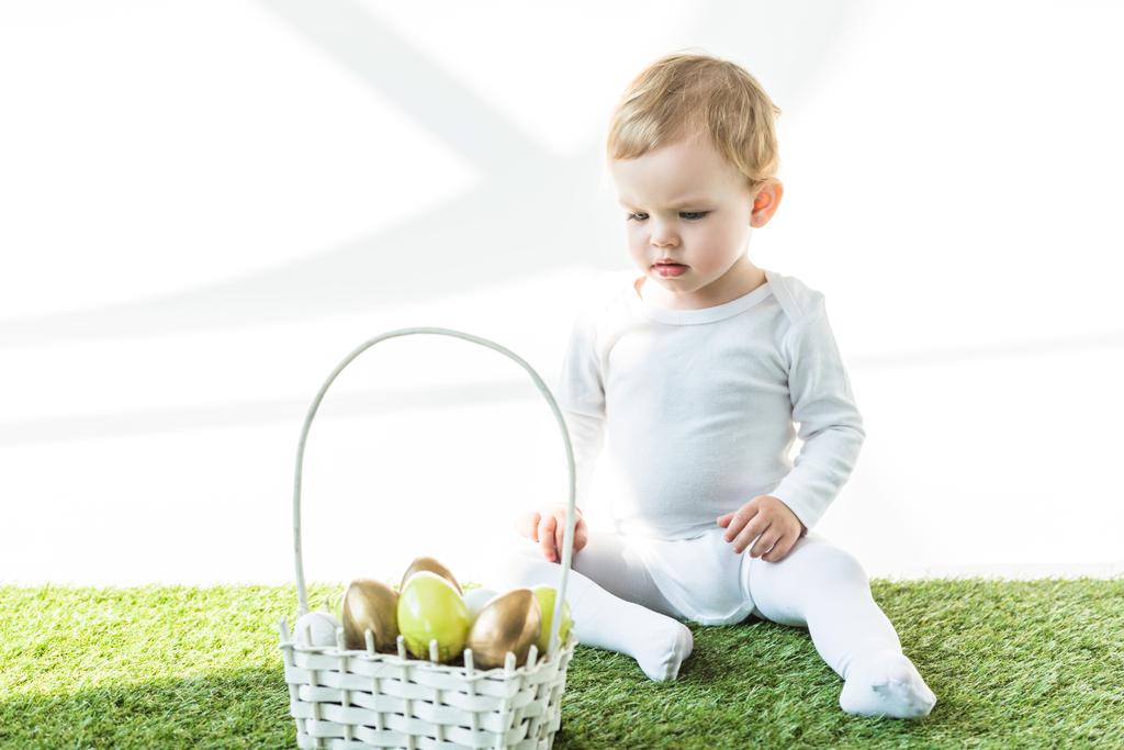 白い上に孤立した黄色と金色のエステルの卵とわらのバスケットの近くに緑の草の上に座っている愛らしい子供 - 写真・画像